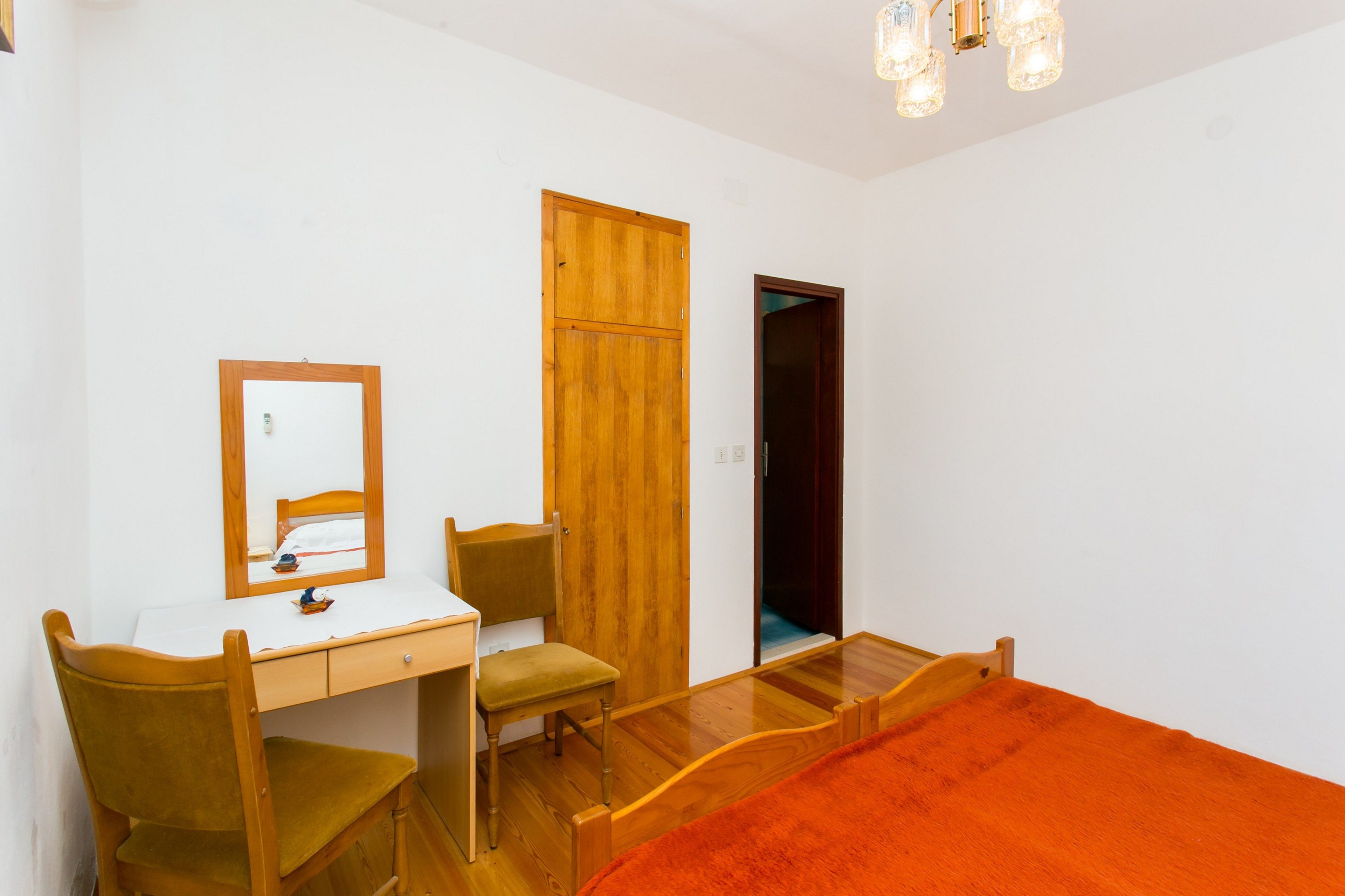 A bedroom with a wooden desk, a chair, a bed with an orange blanket, a wardrobe, and a mirror.