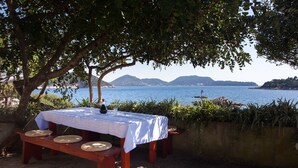 A wooden table with a white tablecloth and woven placemats, set up outdoors with a view of a body of water and distant hills. - Guest House Simunovic (Dubrovnik)