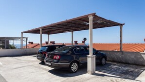 A parking area with a covered structure, two cars, and a view of a red-tiled roof in the background.