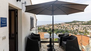 An outdoor balcony with wicker furniture, a large umbrella, and a view of a coastal town.
