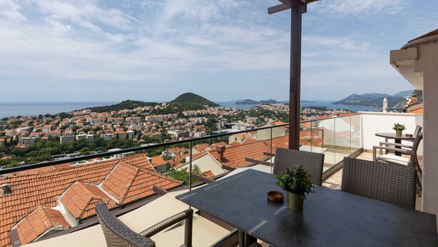 A balcony with a table and chairs, overlooking a coastal town with terracotta rooftops and a view of the sea.