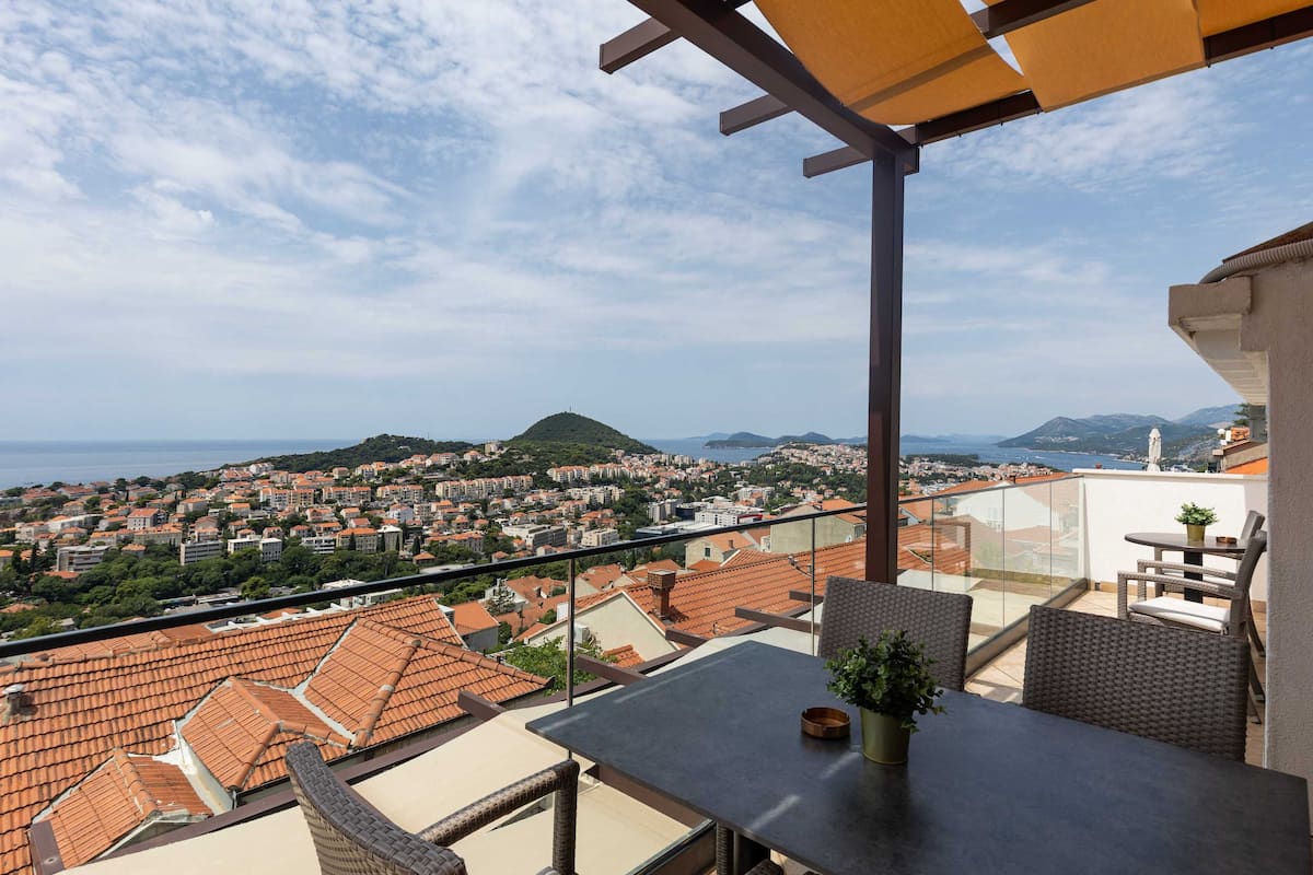 A balcony with a table and chairs, overlooking a coastal town with terracotta rooftops and a view of the sea.