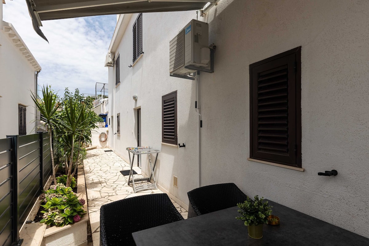 A narrow outdoor patio with stone flooring, a table with chairs, and potted plants.