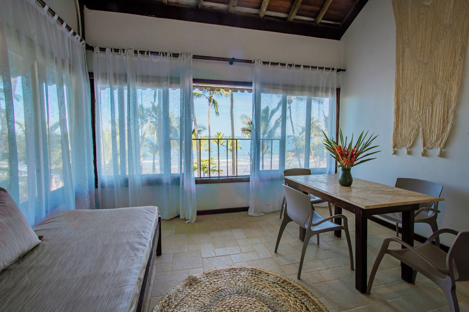 Una habitación con cama, una mesa de comedor con sillas, vistas a la playa y un tapete tejido en el suelo.
