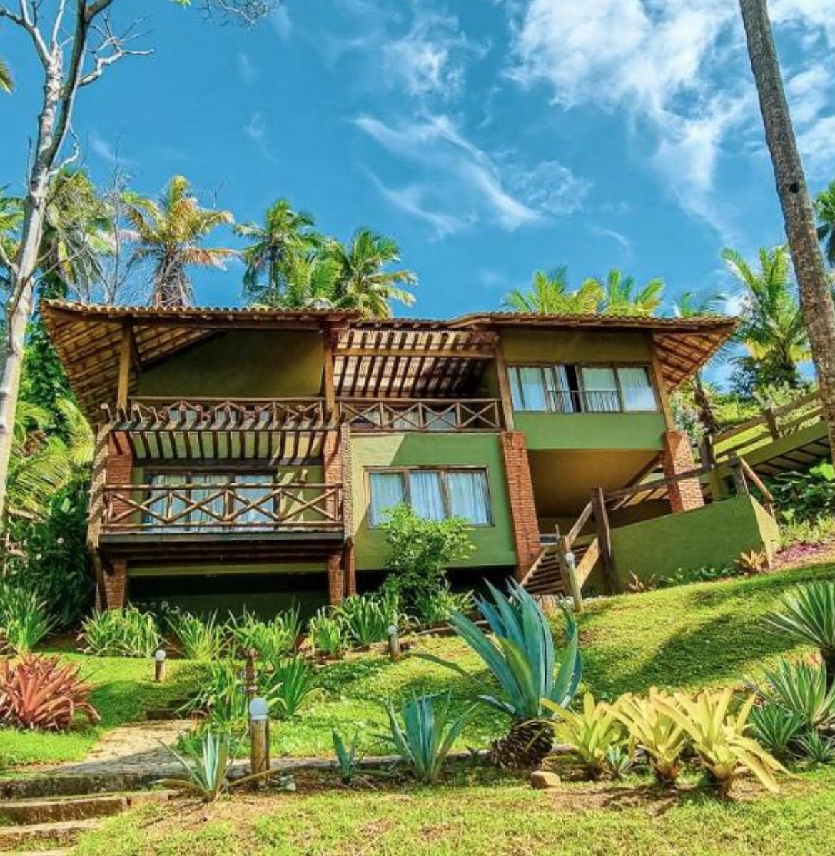 A two-story building with a balcony, surrounded by tropical vegetation.