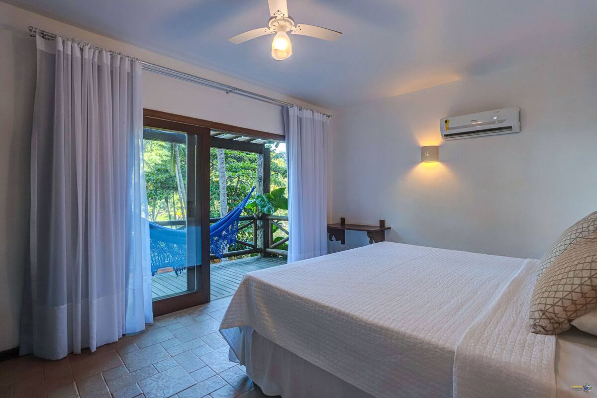 Une chambre avec un lit, des rideaux, un appareil de climatisation et une vue sur un hamac et la verdure à l’extérieur.