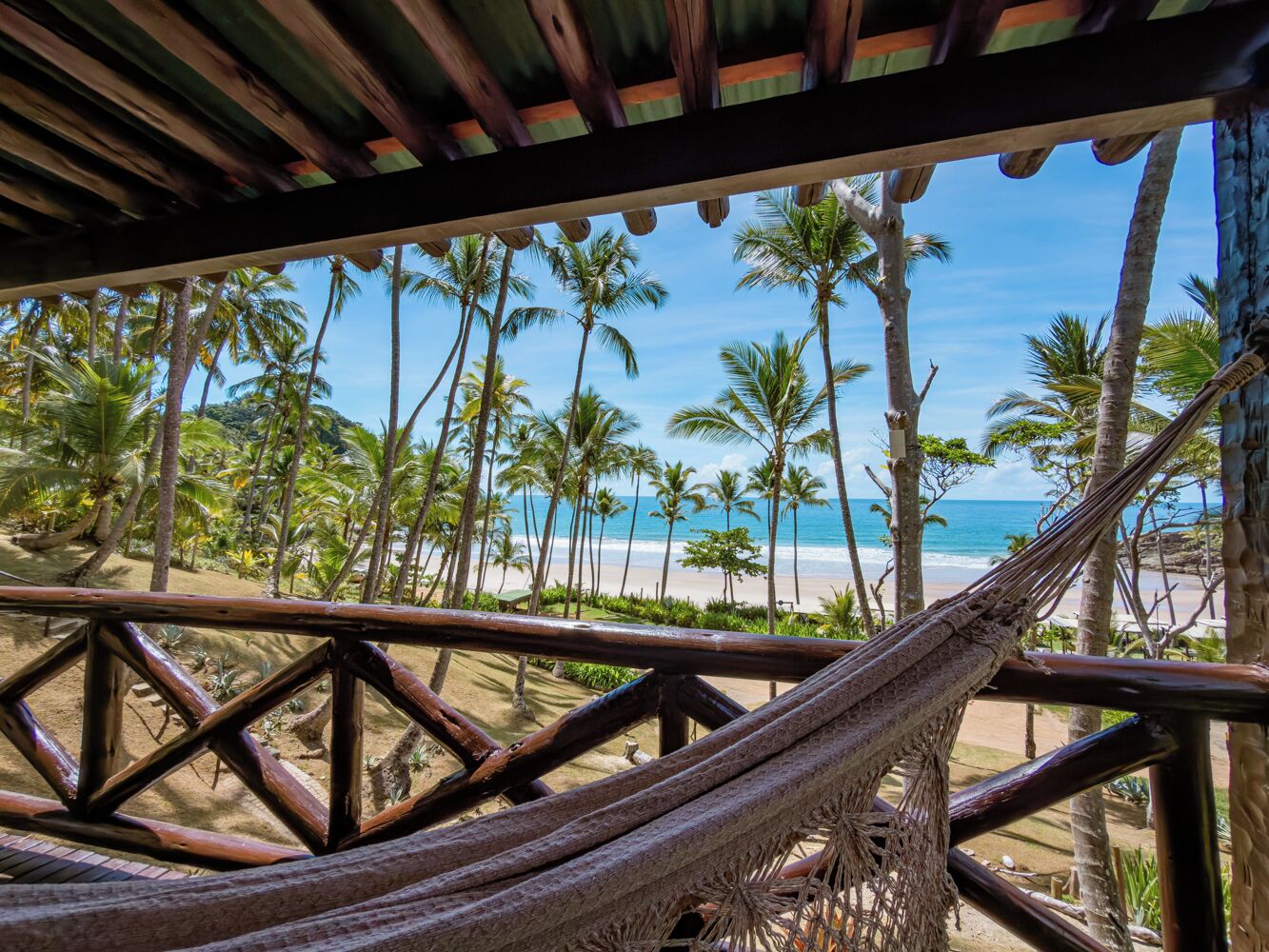 A hammock under a wooden structure with a view of a beach and palm trees.