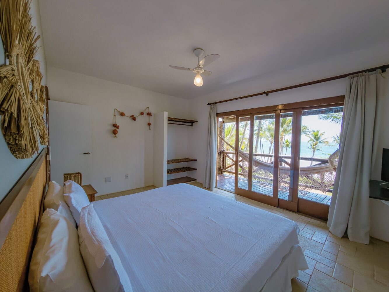 Un dormitorio con una cama grande, ventilador de techo, vista a la playa y balcón con barandilla de madera.