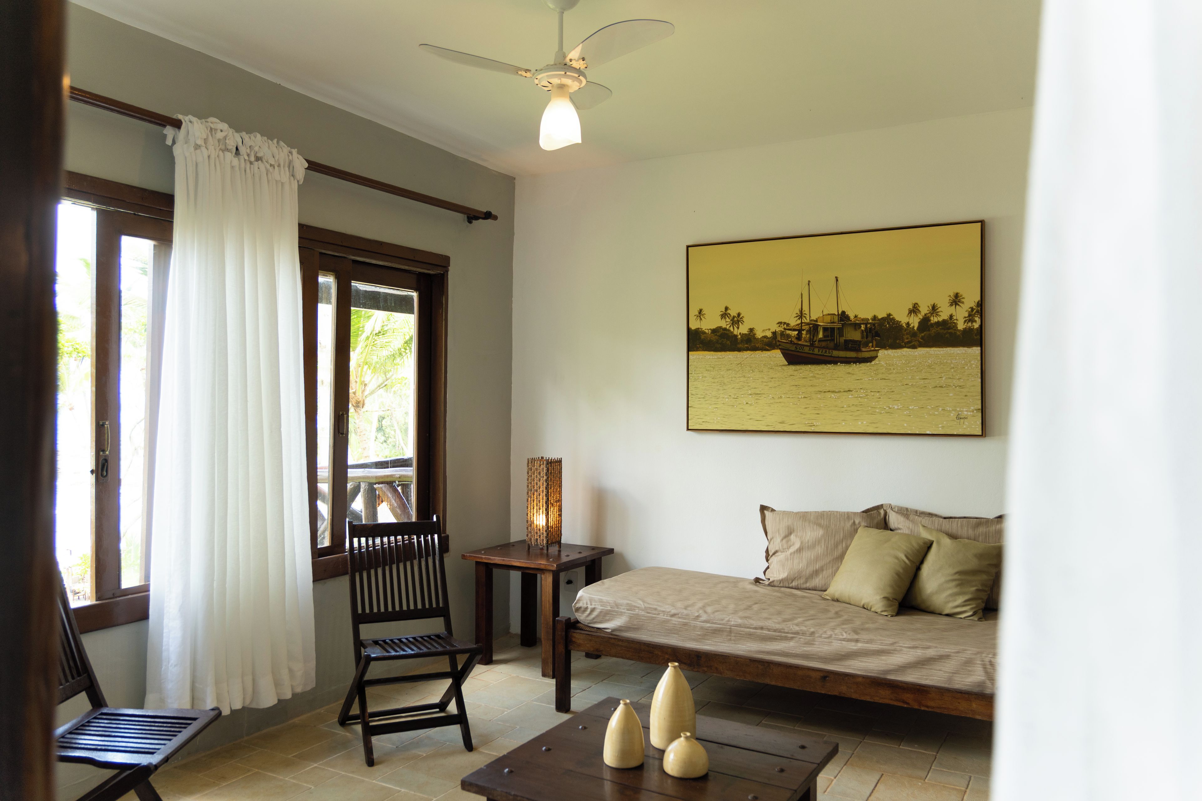Une pièce comprenant un lit, une chaise, une table, un ventilateur de plafond, une fenêtre avec des rideaux et un tableau représentant un bateau accroché au mur.