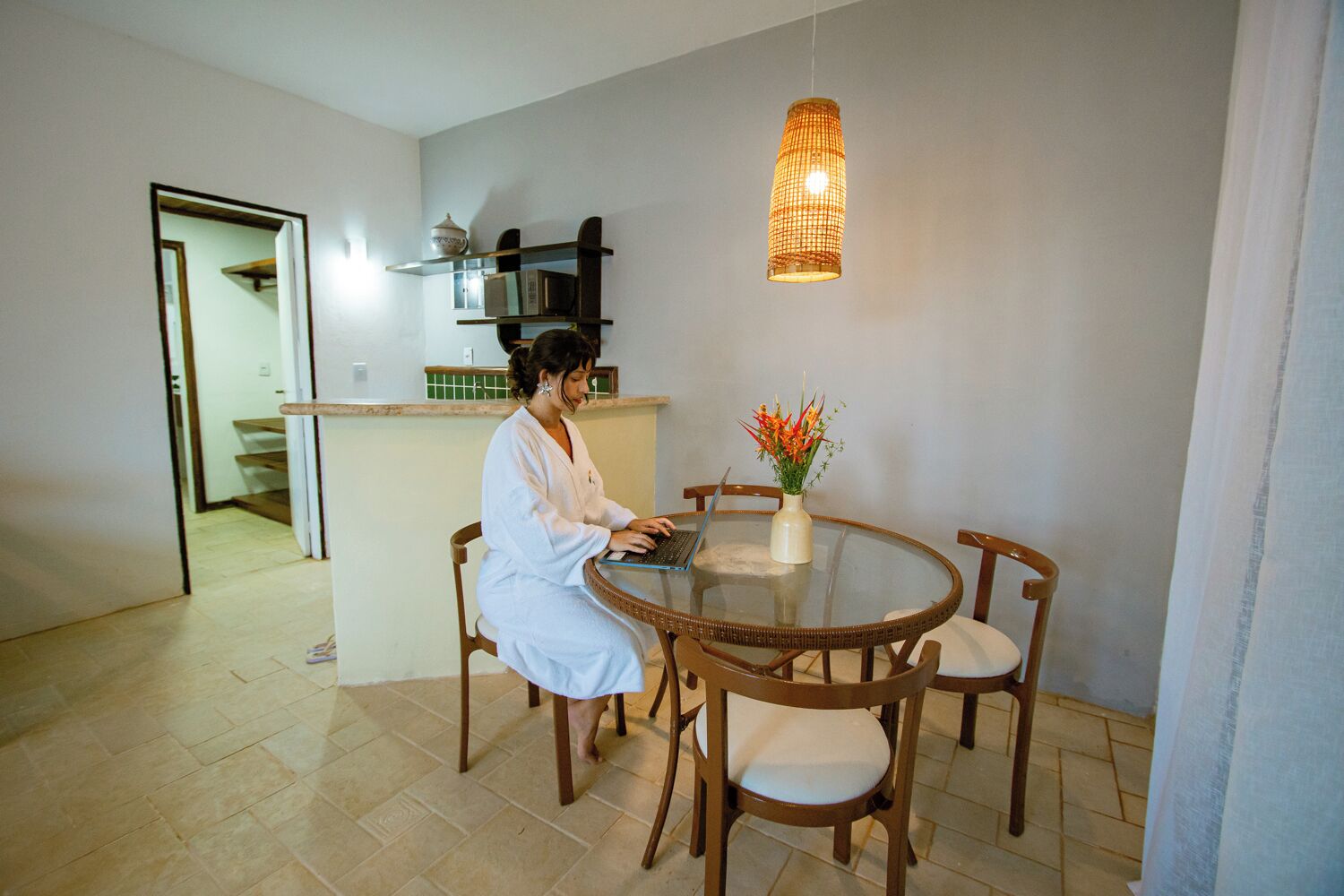 A person seated at a round dining table with a laptop, surrounded by chairs, in a room with a hanging light fixture and a vase with flowers on the table.