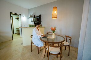 Une personne est assise à une table ronde, devant un ordinateur portable, entourée de chaises, dans une pièce où l’on aperçoit un luminaire suspendu et un vase rempli de fleurs posé sur la table.
