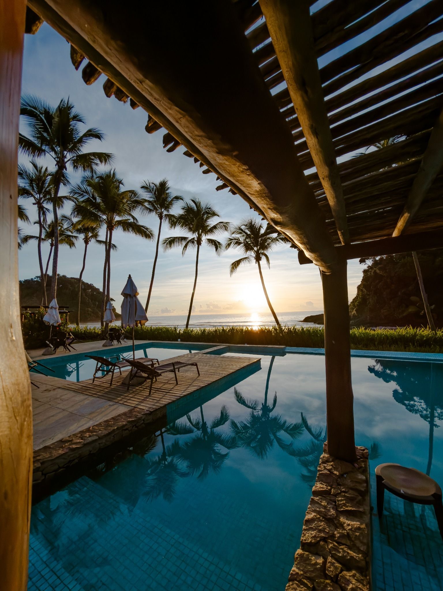 Área de piscina com espreguiçadeiras e guarda-sóis, palmeiras e vista para o pôr do sol.