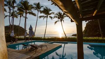 Área de piscina com espreguiçadeiras e guarda-sóis, palmeiras e vista para o pôr do sol.