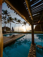 Zona de piscina con sillas de descanso y sombrillas, palmeras y vista al atardecer.
