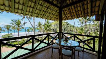 Un espace extérieur aménagé, abrité et doté d’une table et de chaises, offrant une vue sur une plage et des palmiers.