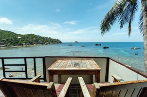 Mesa y sillas de madera en un balcón con vista a una playa donde se ven barcos y una palmera.
