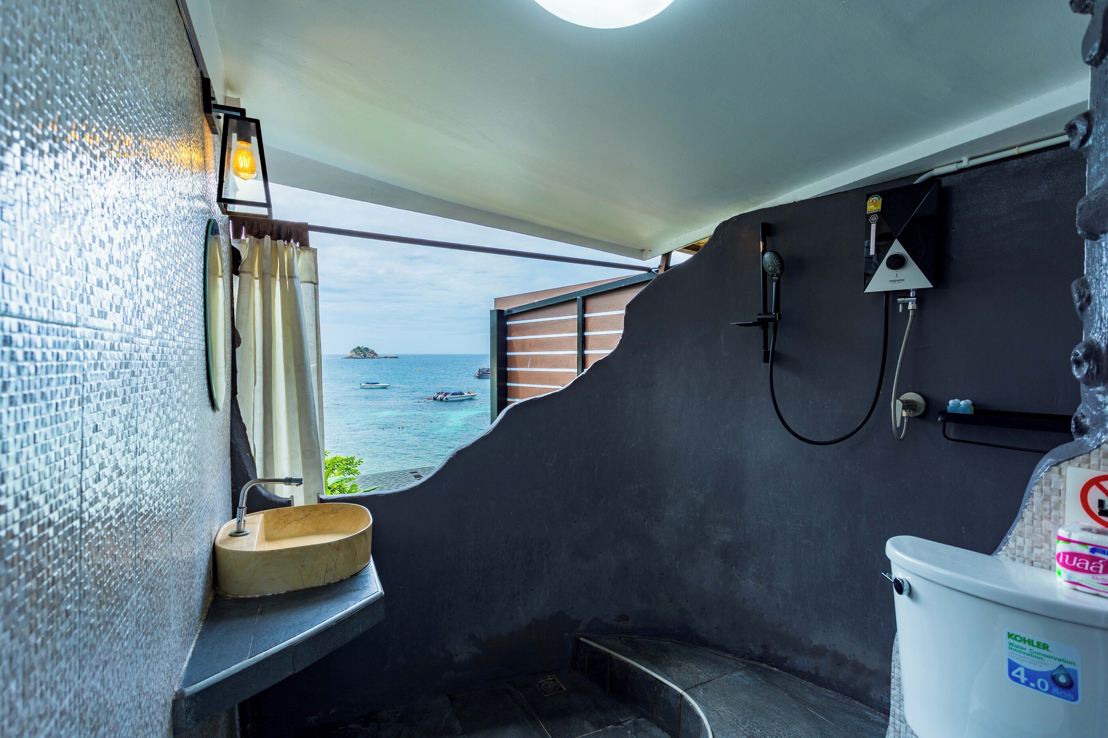 A bathroom with a toilet, a sink, and a shower. There is a view of the sea through the window.