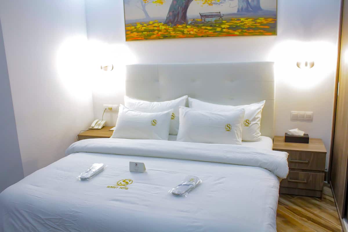 A hotel room with a large bed, white bedding, a painting of a tree and bench, bedside lamps, a phone, and a tissue box on the nightstand.