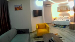 Una sala moderna con un sofá, un sillón amarillo, un televisor de pantalla plana y una cama con edredón blanco.