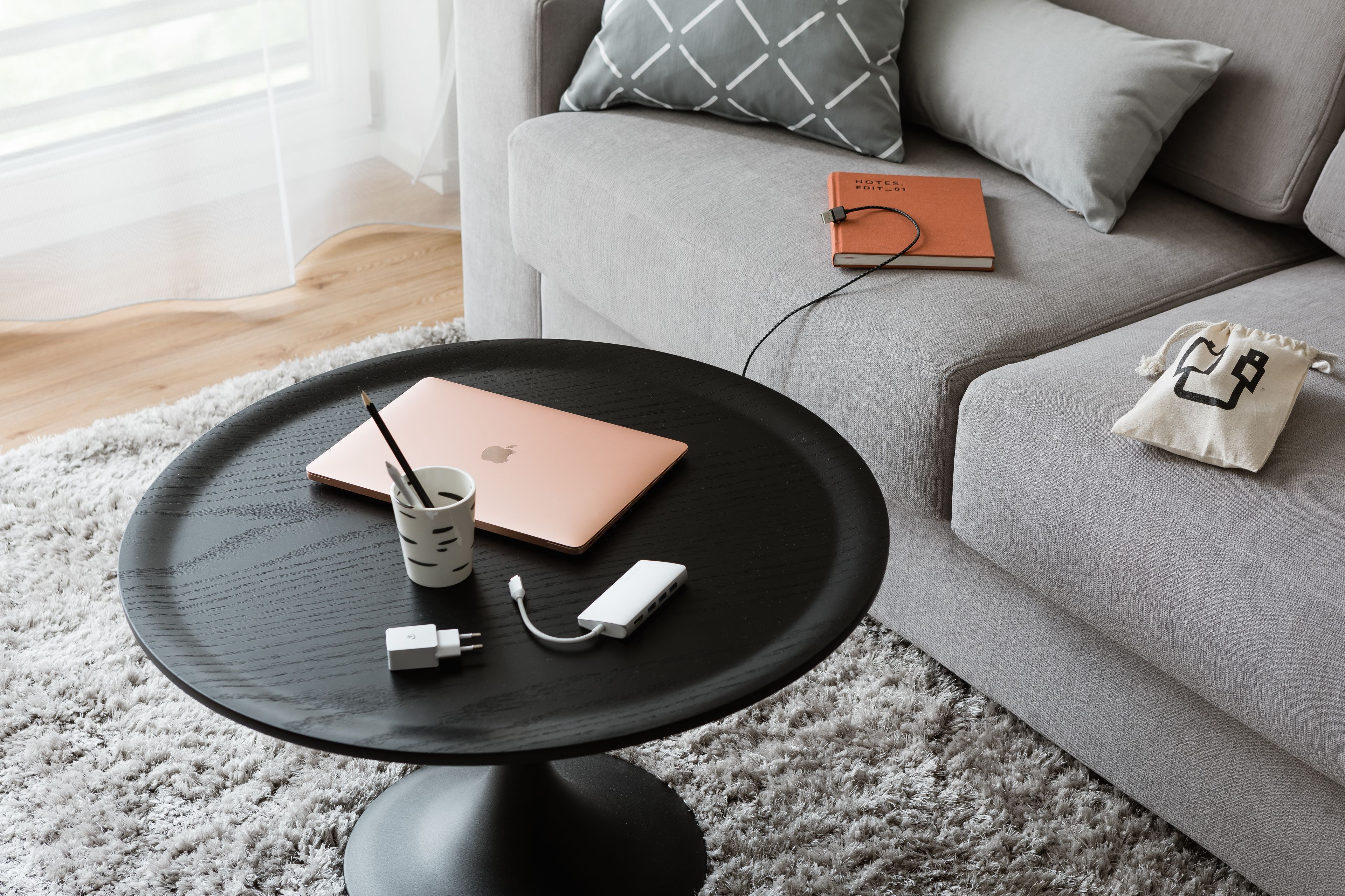 A modern living room with a grey sofa, a round black coffee table, a laptop, a notebook, and a small white bag.