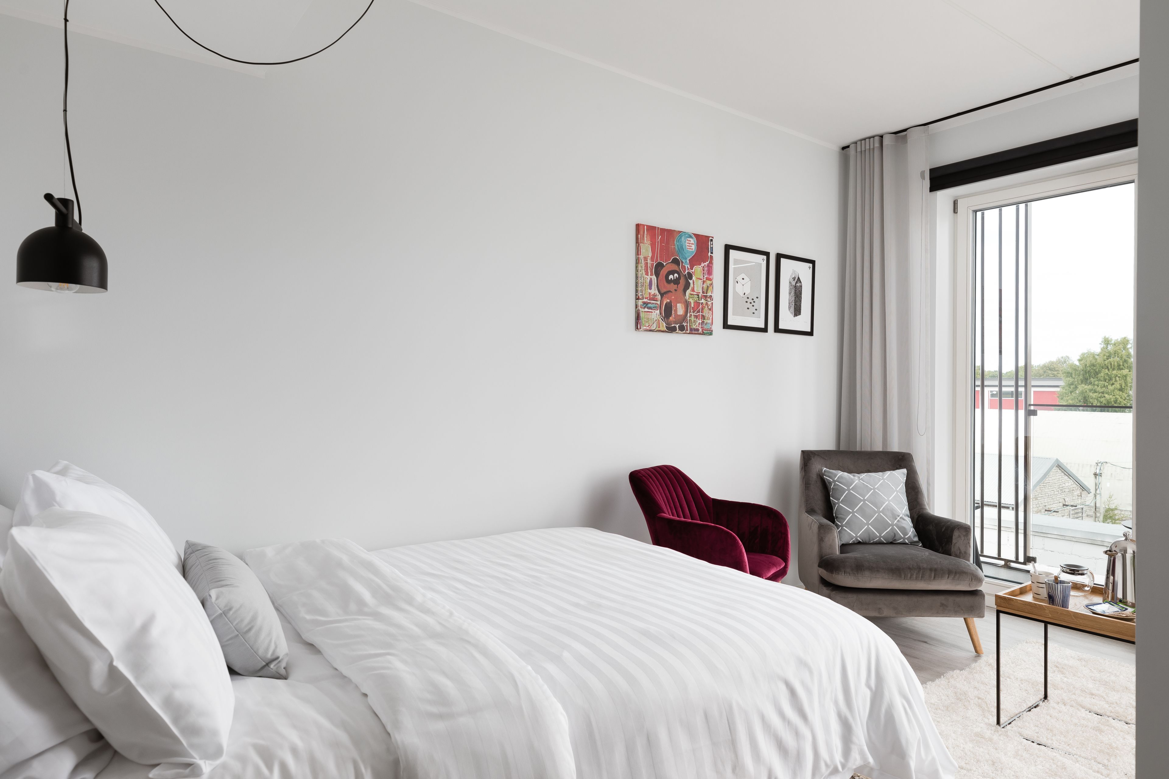 Un dormitorio moderno con una cama grande, una silla roja, un sillón gris y un ventanal con vistas.