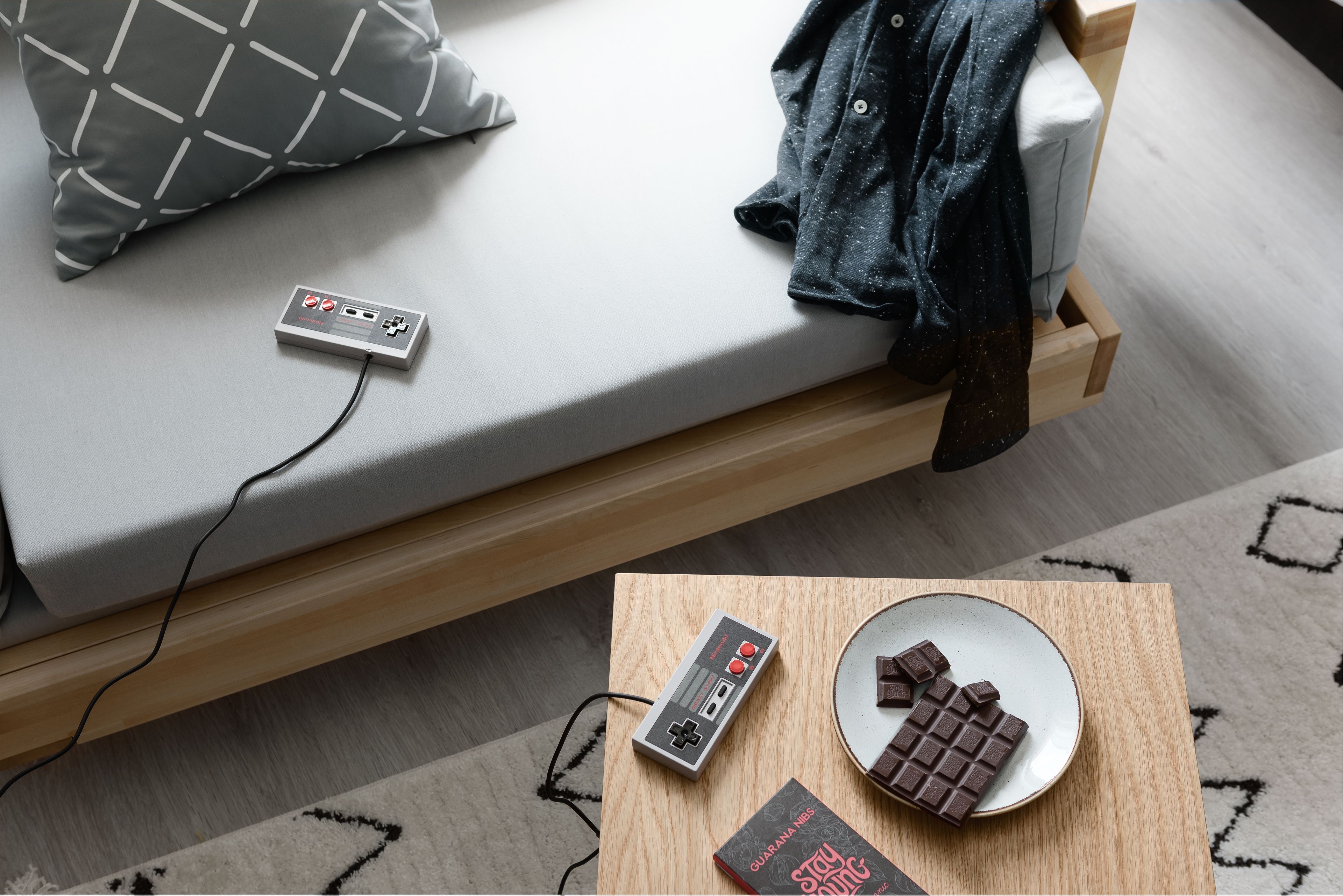 A bed with a gray mattress, a gray pillow with white lines, a denim jacket, a Nintendo 64 console, a plate with chocolate pieces, and a Guarana Nesquik box.