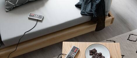 A bed with a gray mattress, a gray pillow with white lines, a denim jacket, a Nintendo 64 console, a plate with chocolate pieces, and a Guarana Nesquik box.