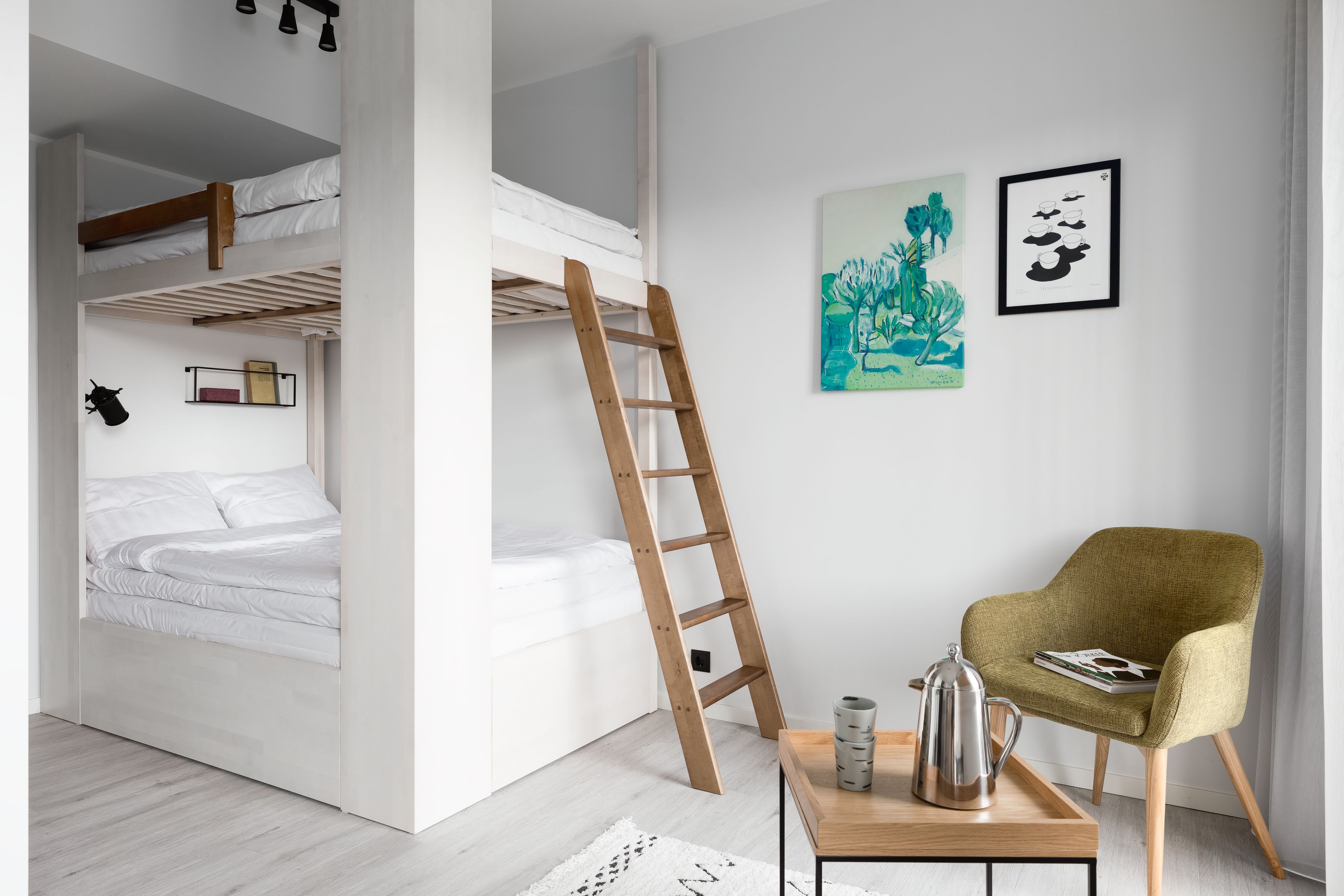 A bedroom with bunk beds, a ladder, a chair, a table with a kettle, and two framed artworks on the wall.