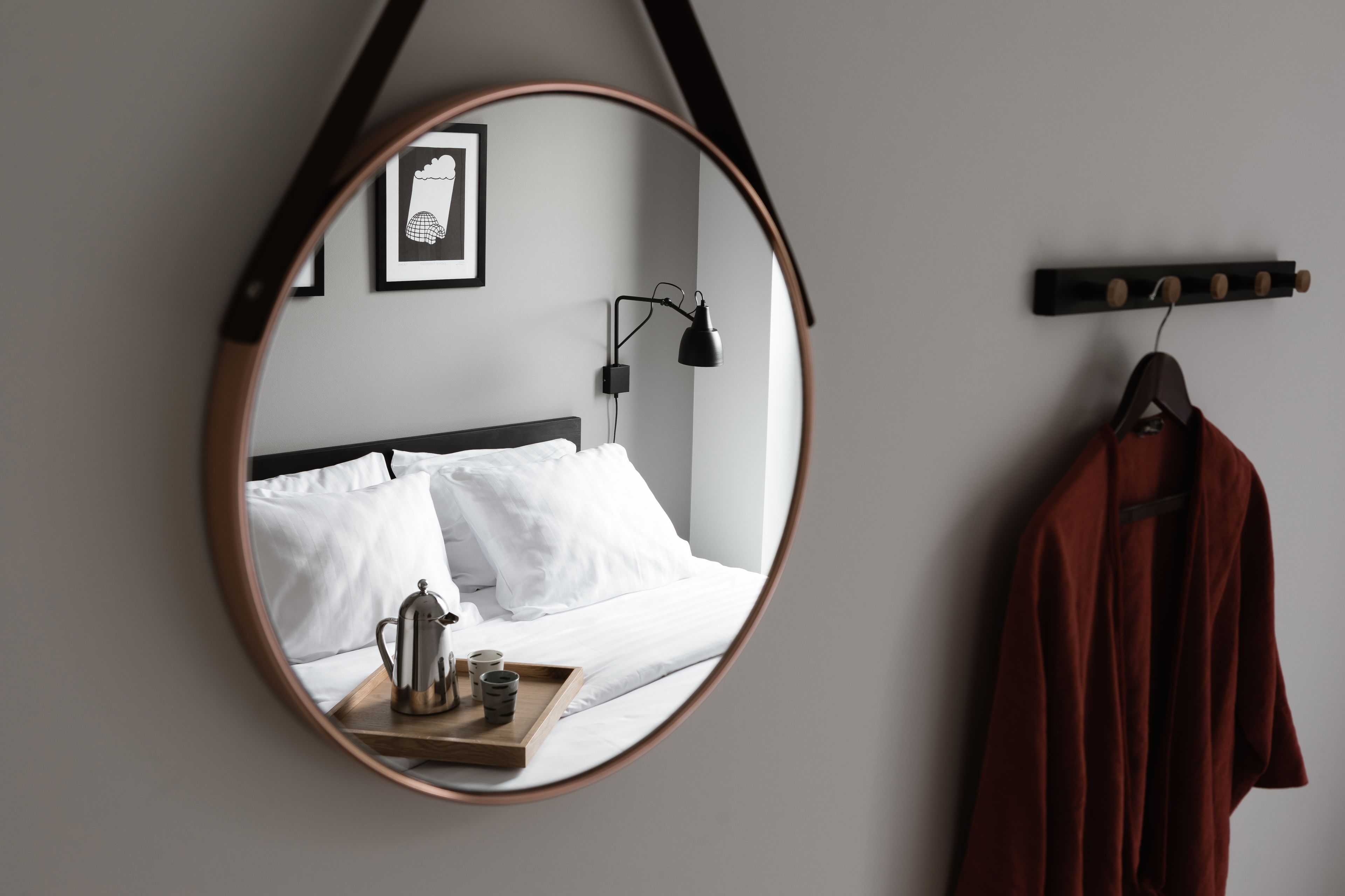 Un dormitorio con un espejo redondo, una cama con ropa de cama blanca, una mesita de noche con tetera y tazas, una bata colgada y una lámpara fijada a la pared.