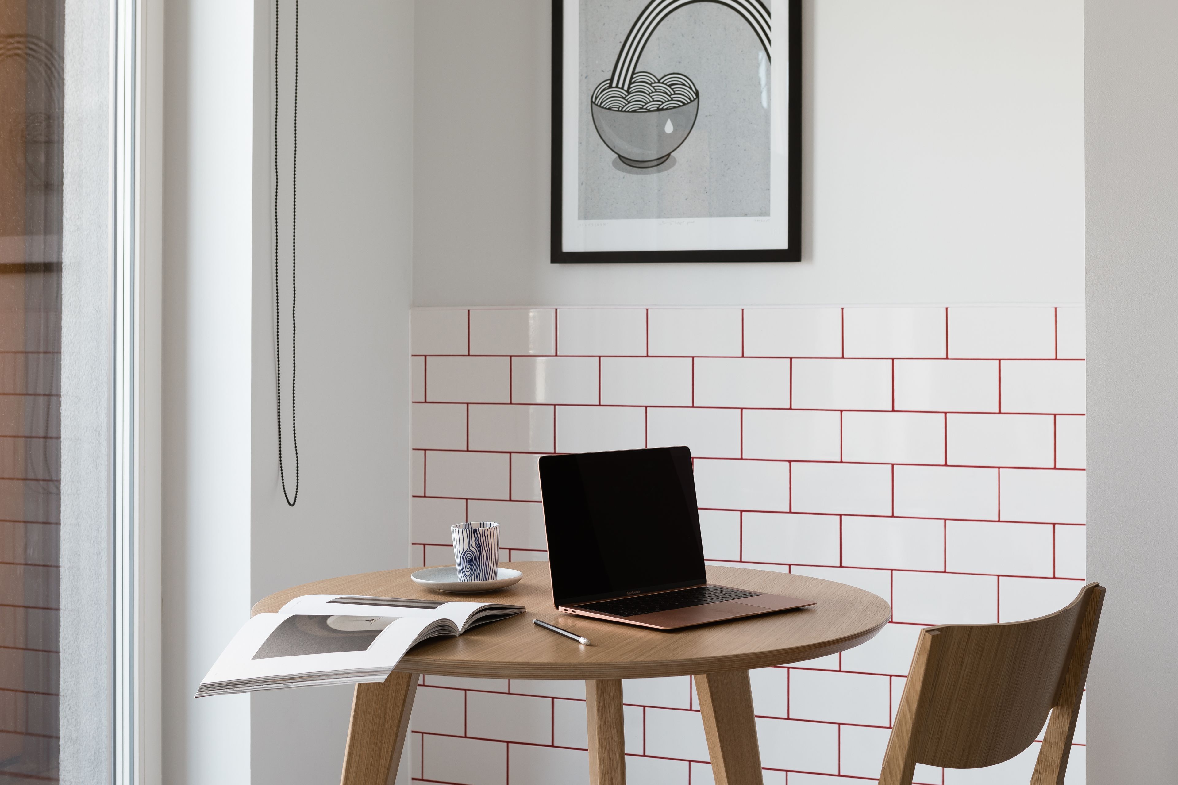 A laptop on a round wooden table with a magazine, a pen, and a glass of water. A framed picture hangs on the wall above the table.