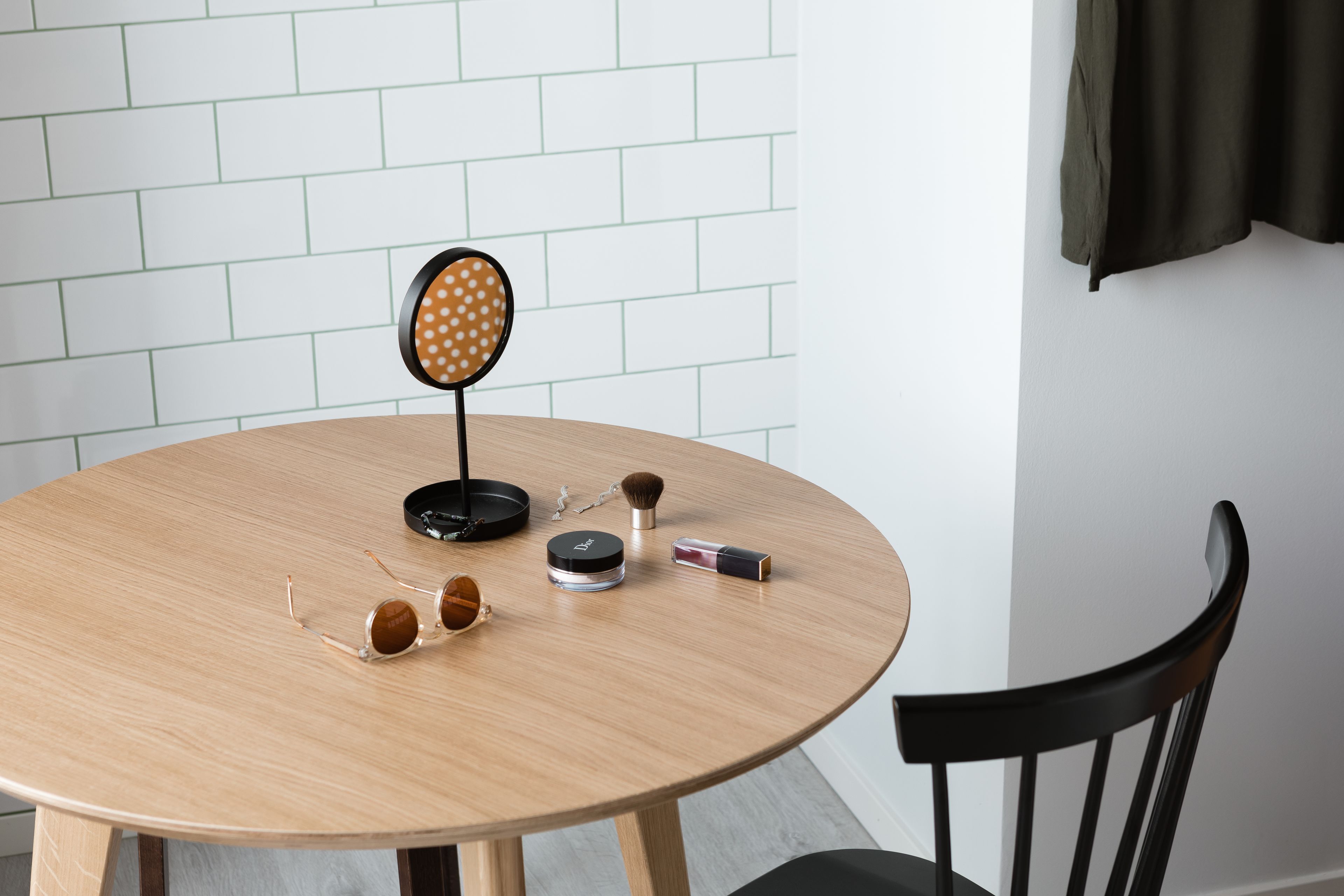 A round wooden table with a black chair, a round mirror with a polka dot frame, and various personal items on the table.
