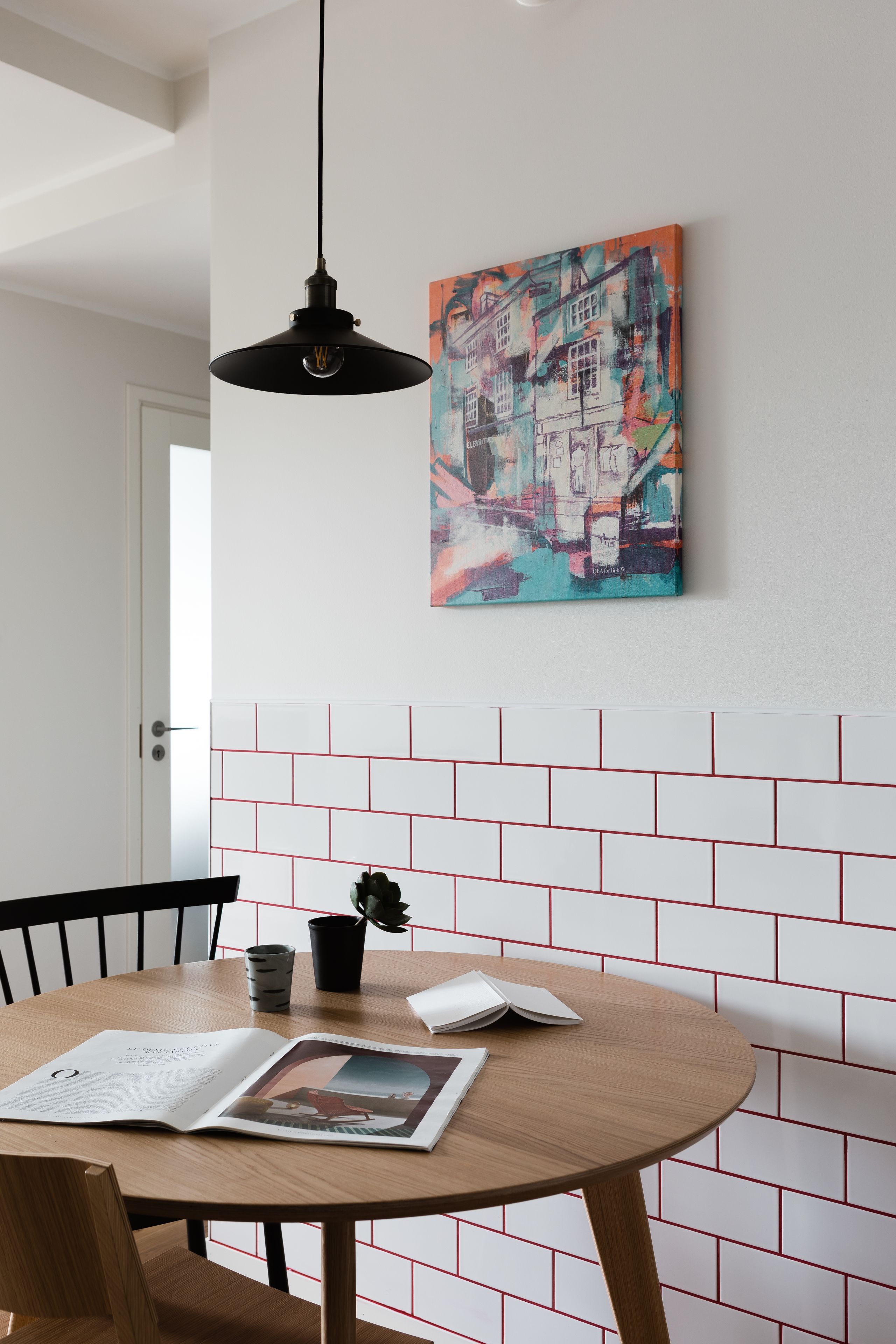 Ein Essbereich mit einem runden Holztisch, einer schwarzen Pendelleuchte und einem farbenfrohen, abstrakten Gemälde an der Wand.