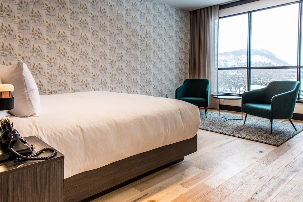 Ein modernes Hotelzimmer mit einem großen Bett, zwei Sesseln, einem kleinen Tisch und Blick auf einen verschneiten Berg durch große Fenster.
