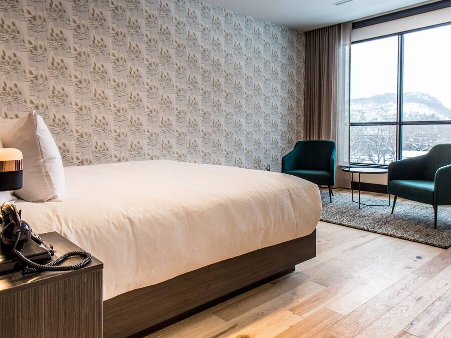 A modern hotel room with a large bed, two chairs, a small table, and a view of a snowy mountain through large windows.