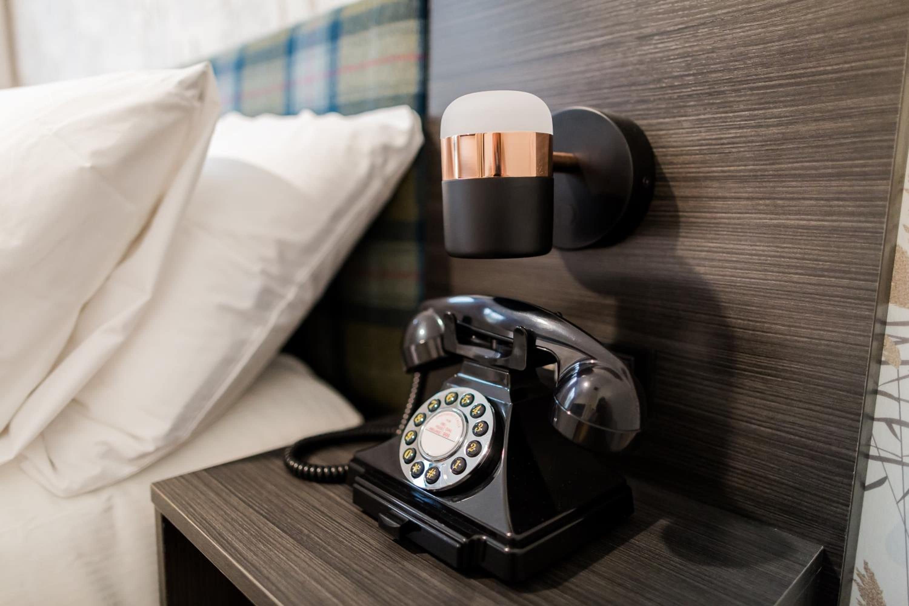 A black rotary telephone on a bedside table with a pillow and a wall-mounted telephone receiver.