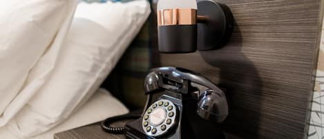 A black rotary telephone on a bedside table with a pillow and a wall-mounted telephone receiver.