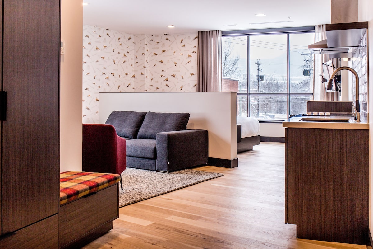 Ein modernes Hotelzimmer mit einer Sitzecke, einer kleinen Küchenzeile und einem großen Fenster mit Blick auf die Stadt.