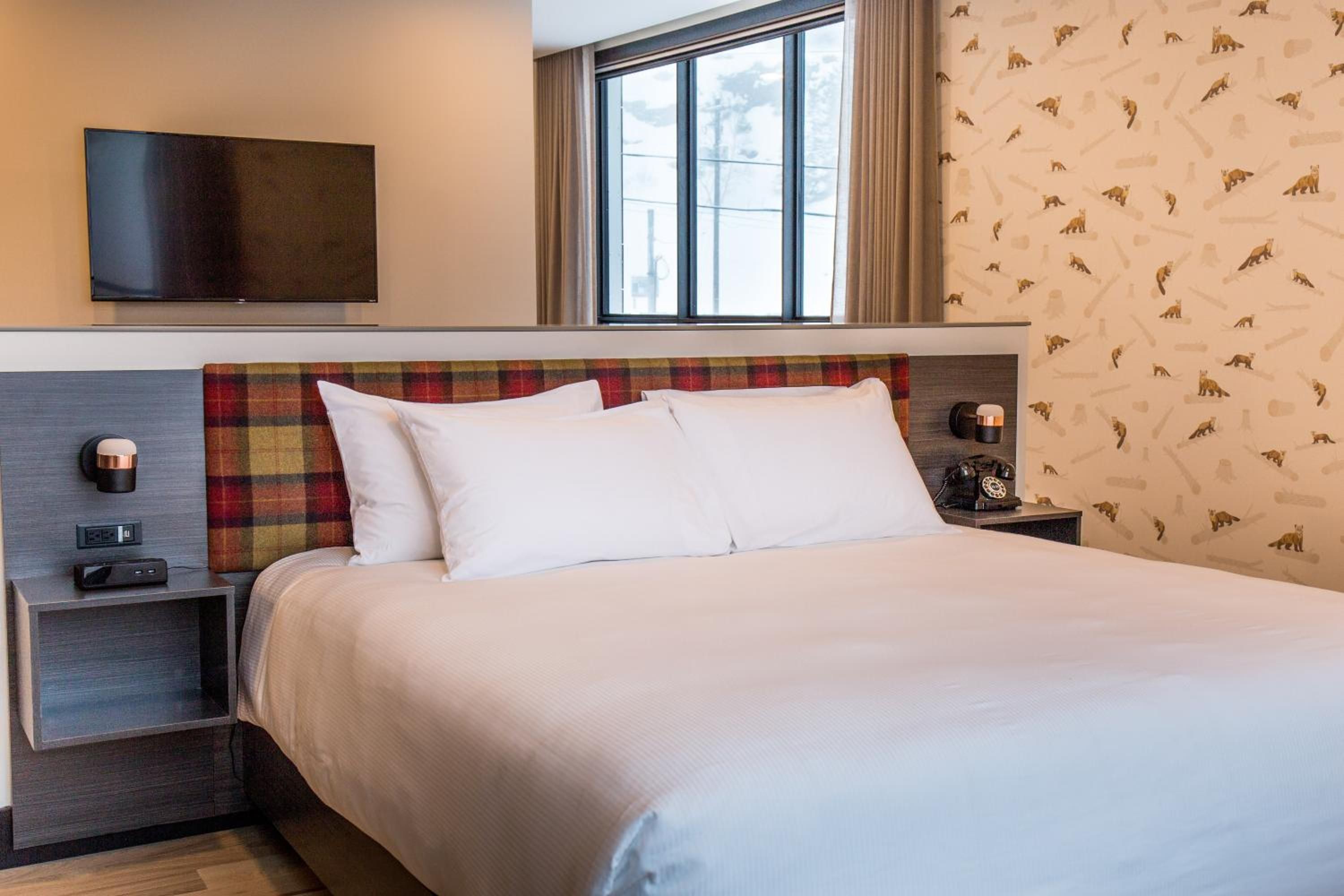 A hotel room with a bed, a TV mounted on the wall, a nightstand, and a window with curtains.
