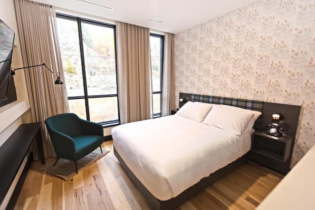 A hotel room with a bed, a chair, a nightstand, and a large window.