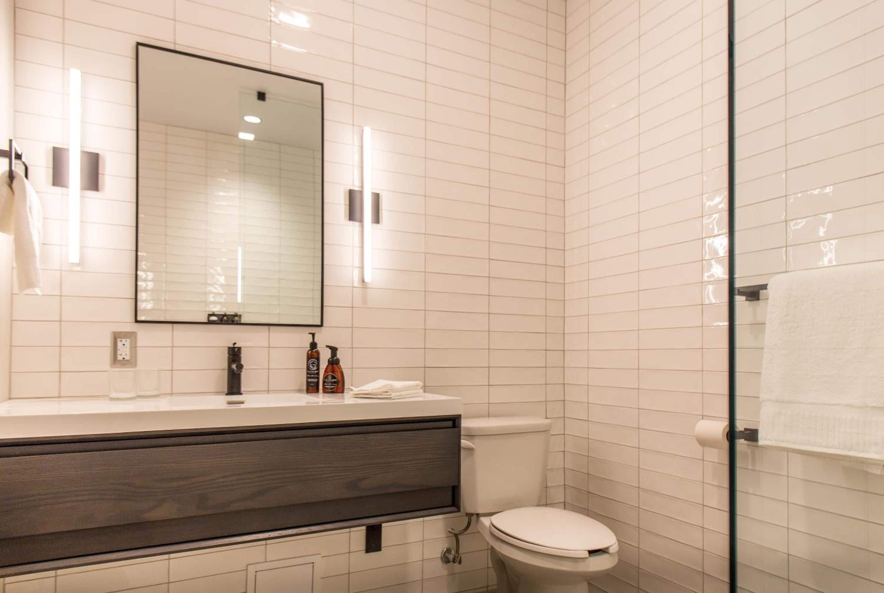 Une salle de bain moderne avec un mur recouvert de carreaux blancs, un grand miroir, un meuble-lavabo en bois foncé, des toilettes et une cabine de douche en verre.