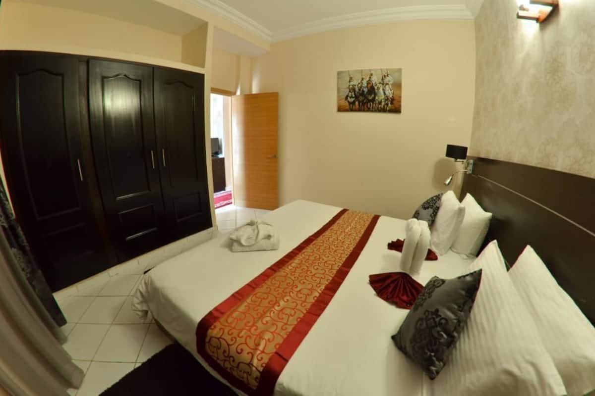 A hotel room with a large bed, a wardrobe, a painting on the wall, and a bathroom with a mirror and towel rack.
