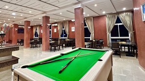 A pool table in a well-lit indoor area with multiple tables and chairs, suggesting a recreational space within a hotel. - Motel Al Akha (Ain Kansra)