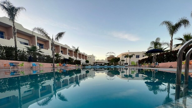 A clear swimming pool with a tiled edge, surrounded by palm trees and hotel buildings. - Motel Al Akha (Ain Kansra)