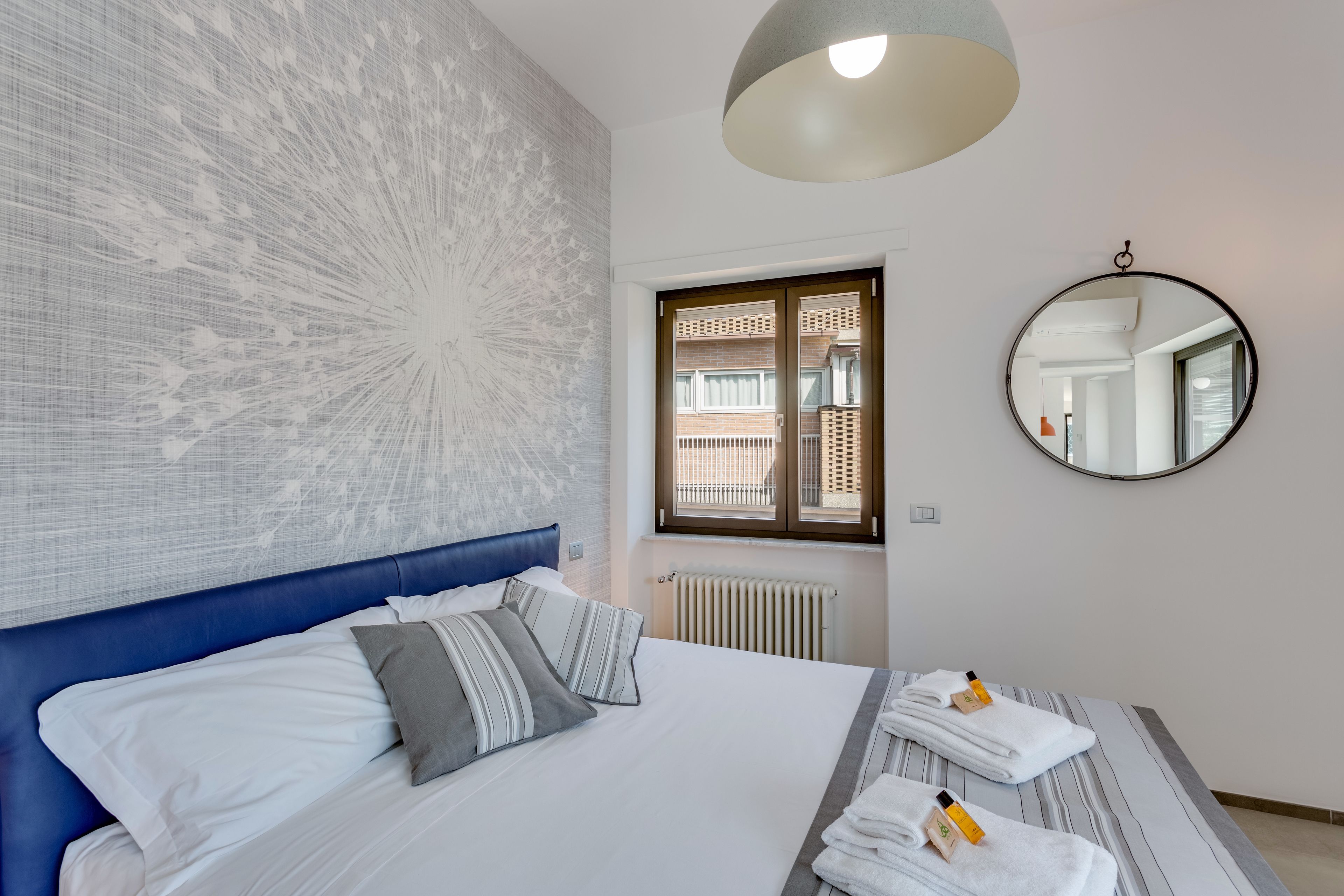 A hotel room with a large bed, a blue headboard, a round mirror, and a view of the outside through a window.