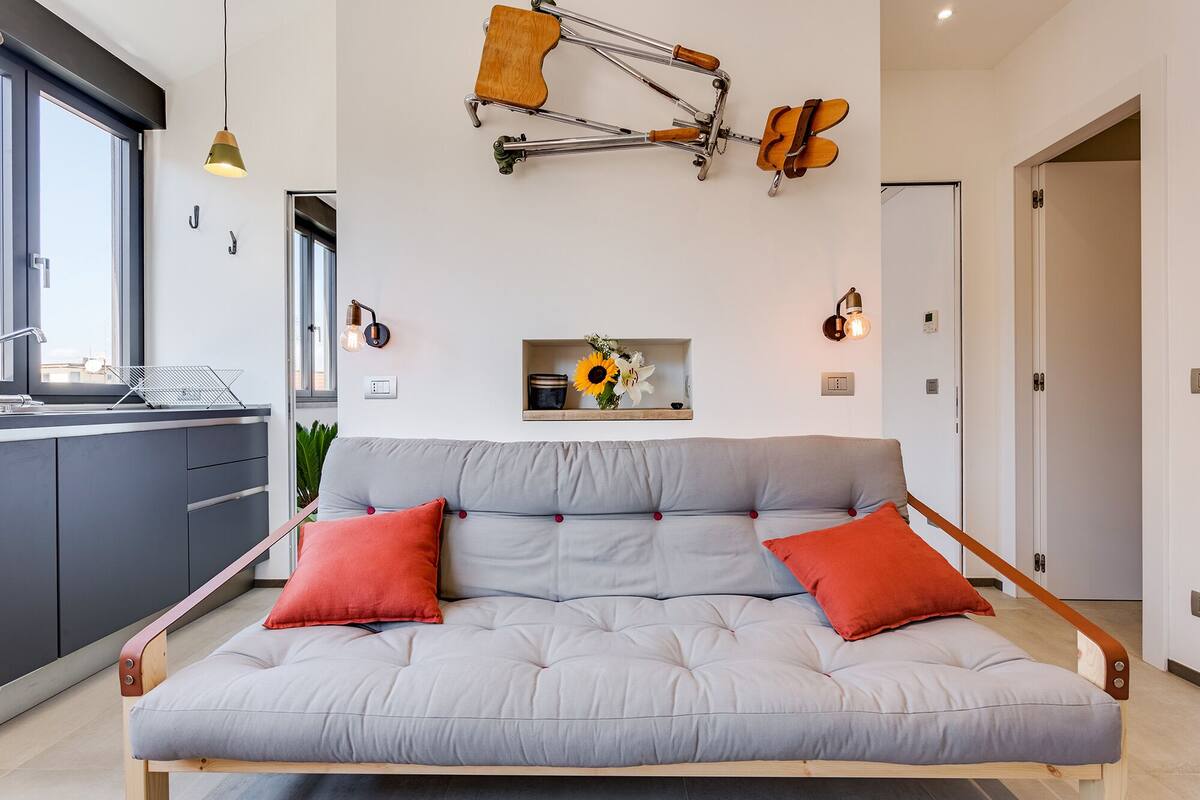 Ein modernes Wohnzimmer mit grauem Sofa, roten Kissen, einer Wanddekoration aus einer Holzgitarre und einem kleinen Regal mit Blumen.