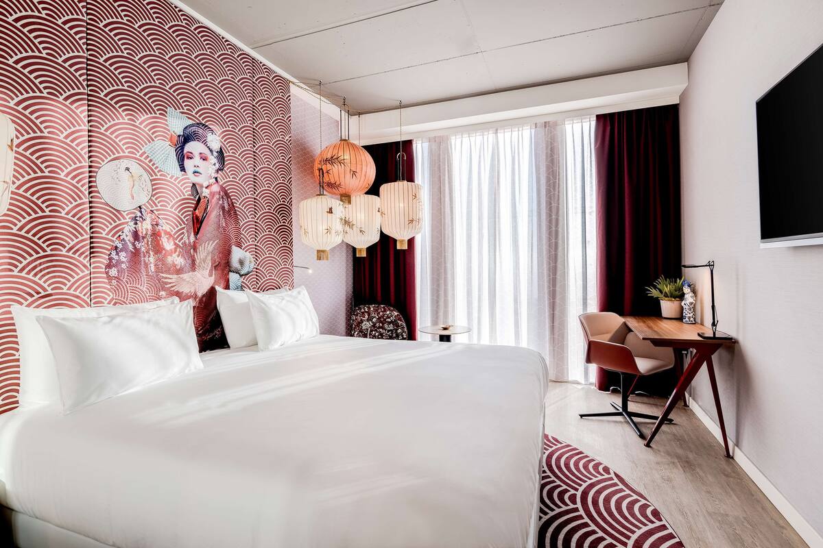 A hotel room with a large bed, a desk with a chair, and a view of the outdoors through a window with curtains.