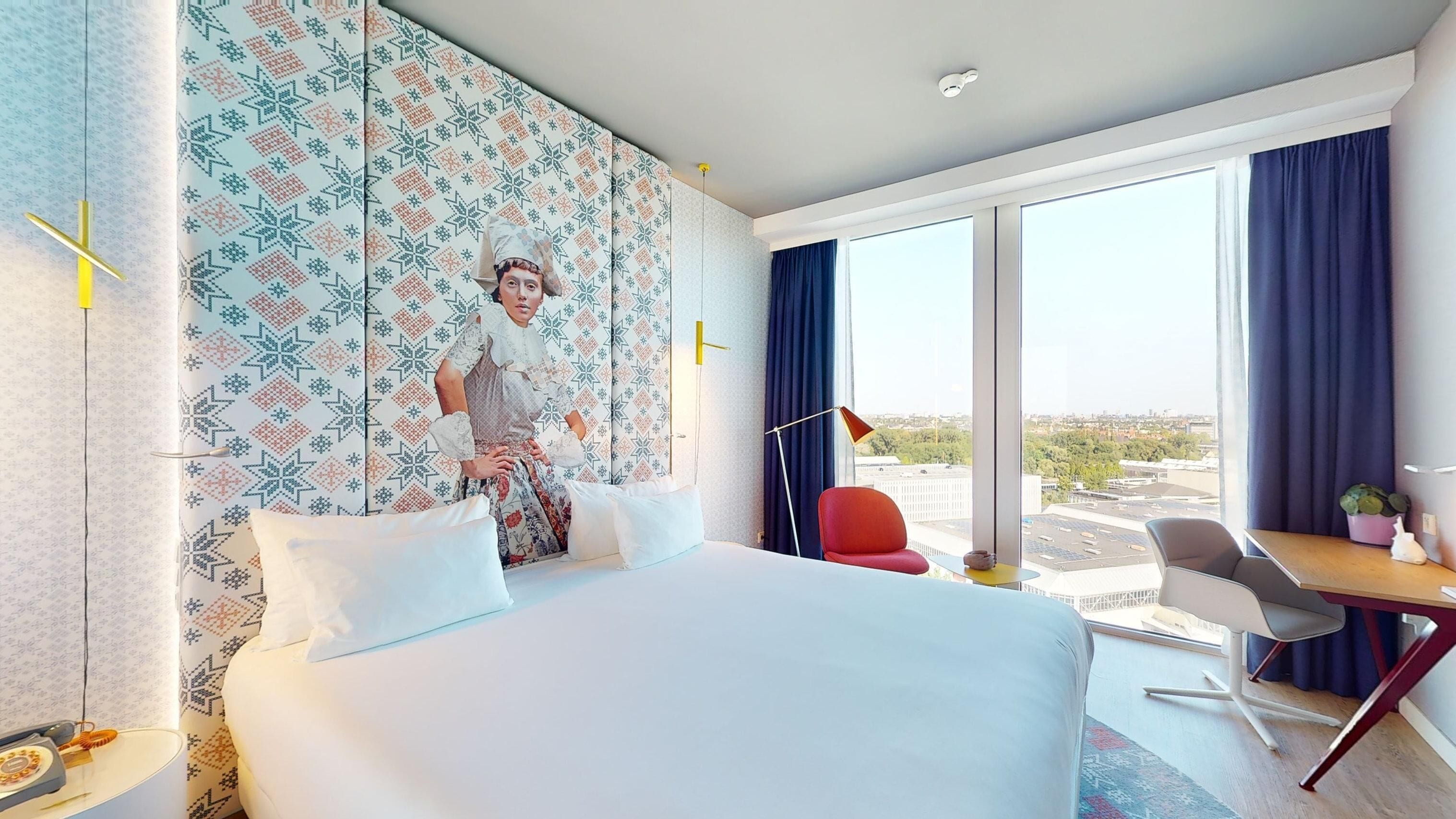 Habitación de hotel con una cama grande, un escritorio, una silla y vistas a la ciudad a través de amplias ventanas.