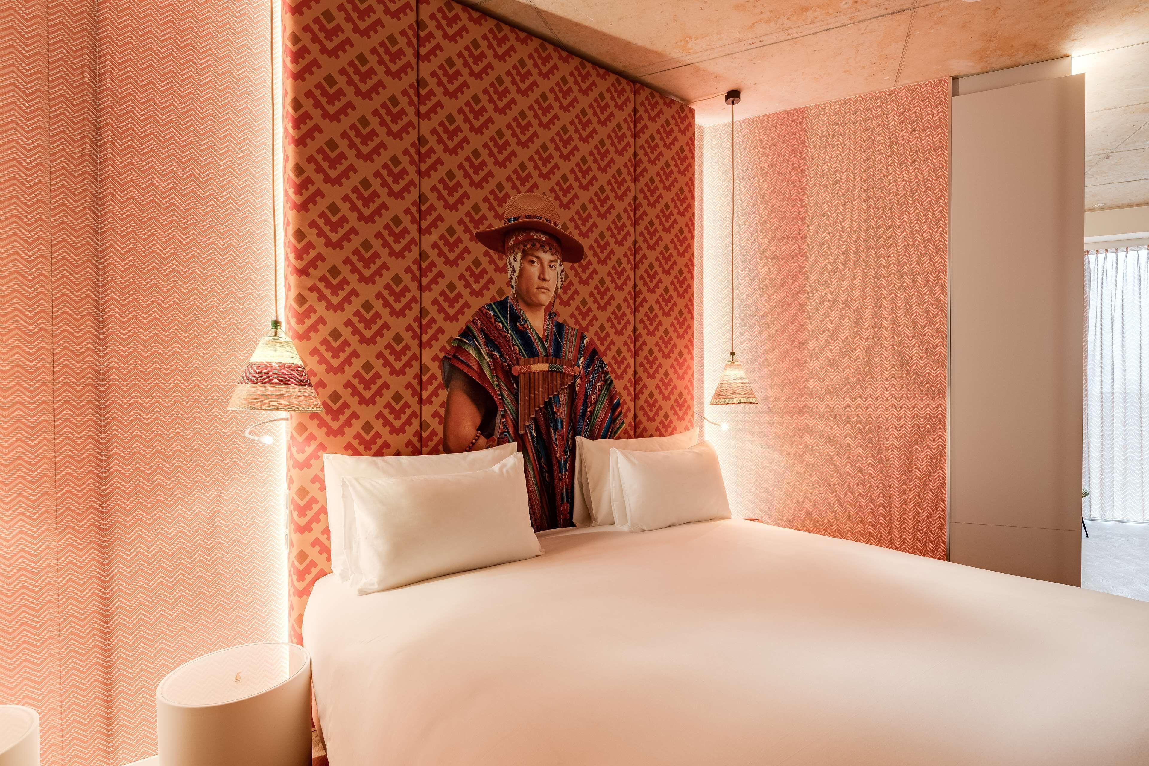 A hotel room with a bed, two bedside lamps, a patterned wall, and a door.