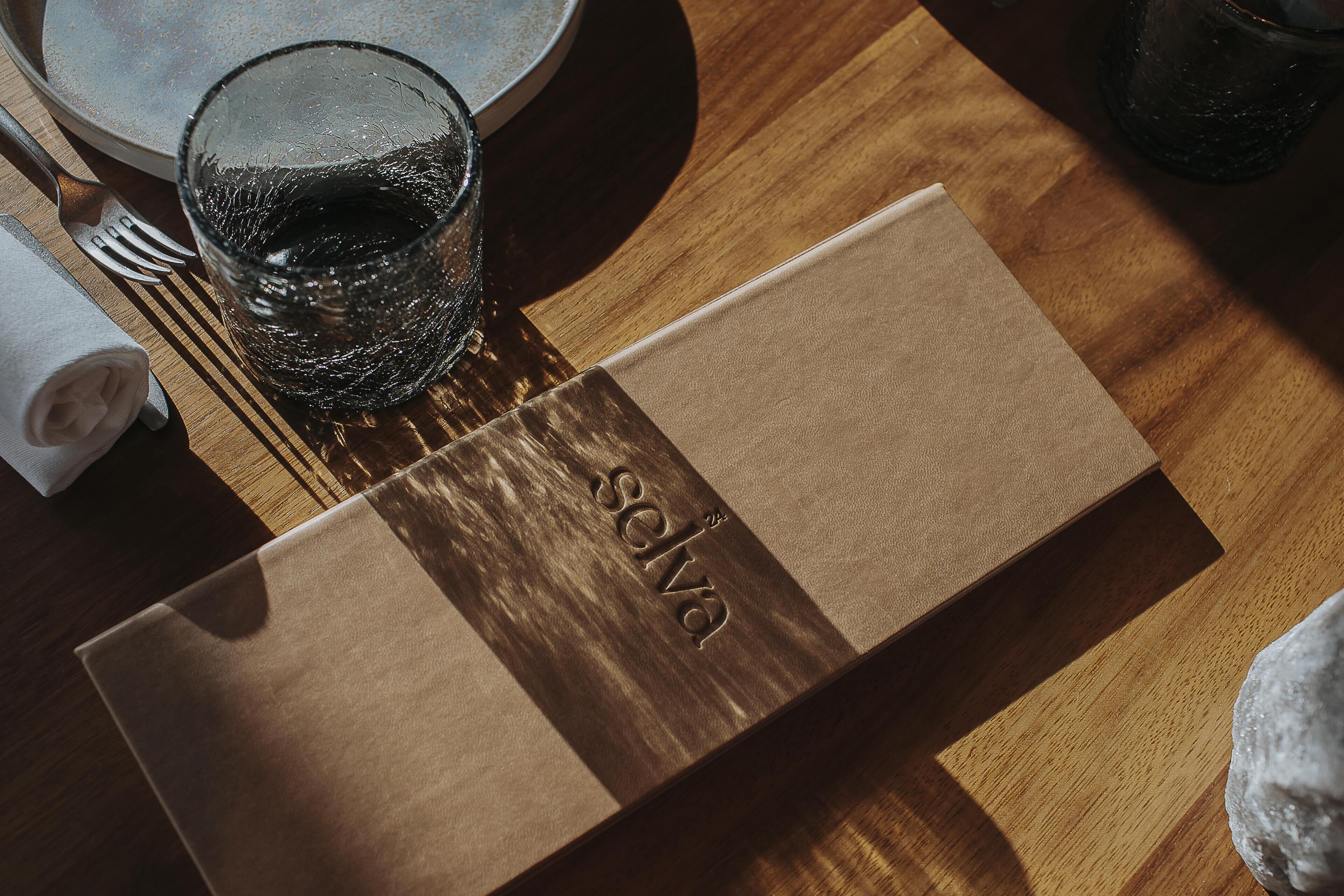 Une table en bois, avec un verre, une serviette pliée, une fourchette et une boîte rectangulaire portant le mot « selva » en relief.