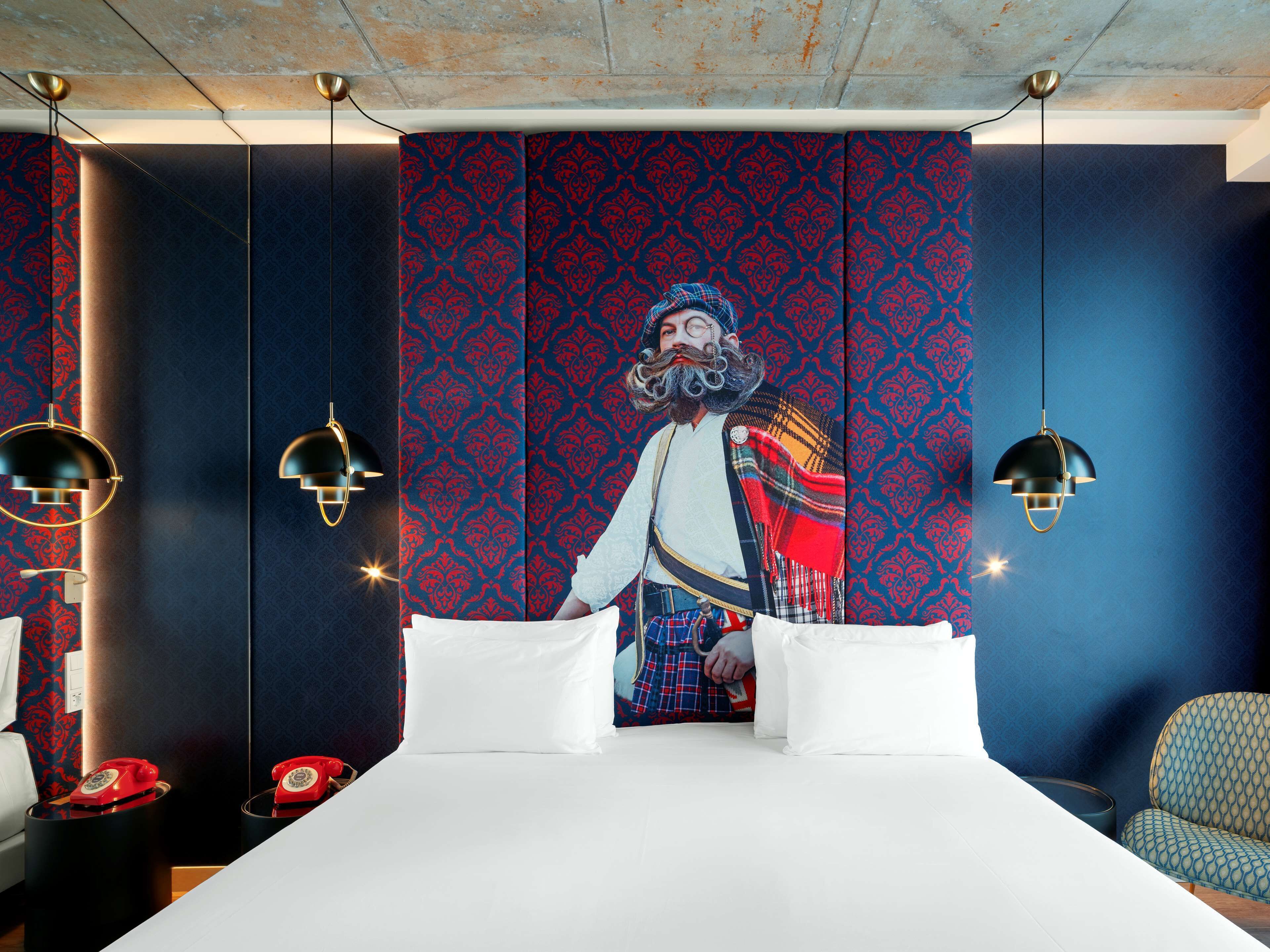 Habitación de hotel con una cama grande, dos mesitas de noche con teléfonos giratorios rojos y una pared de diseño azulado.